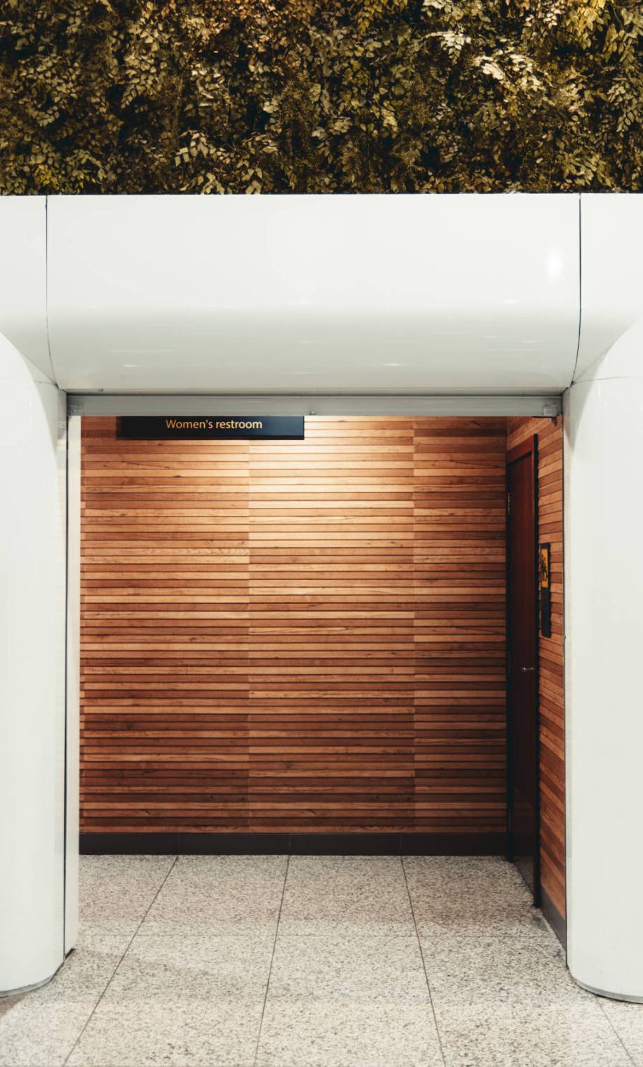 Entrance to a women's restroom with modern design, featuring a white portal, wooden walls, and a vertical garden on the side. Bright, clean, and accessible